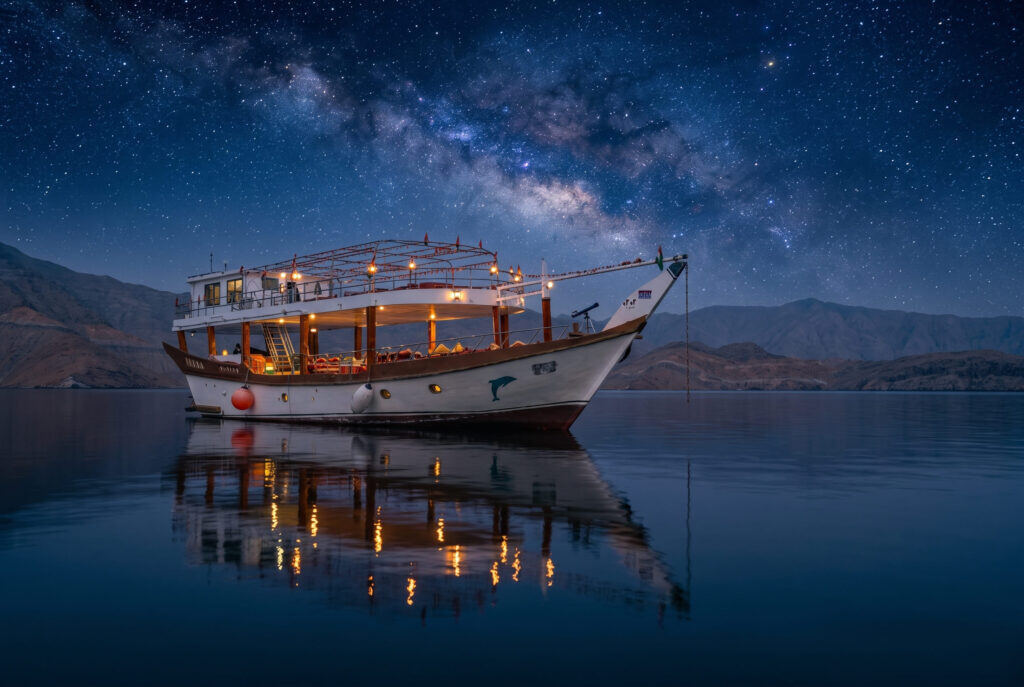 Traditional dhow anchored in a quiet Musandam fjord at dusk with deck lantern lighting and sleeping cushions